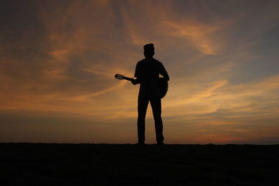 Rear view of silhouette man with guitar standing against sky during sunset