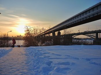 Bridge over snow covered field against sky during sunset