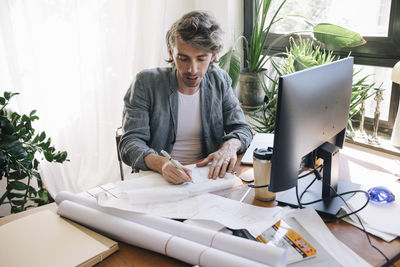 Confident male architect sketching blueprint on paper at desk in home office