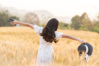 Back of long hair woman in white dress stretching her arms holding her hat and phone in barley field