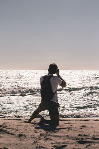 Rear view of man kneeling at beach against clear sky during sunset