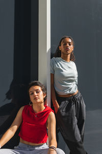 Serious multiracial female friends in casual clothes looking at camera while standing near gray wall of building on sunny street