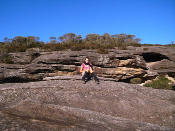 Full length of woman sitting on rock against clear blue sky