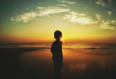 Silhouette boy standing at beach against cloudy sky during sunset