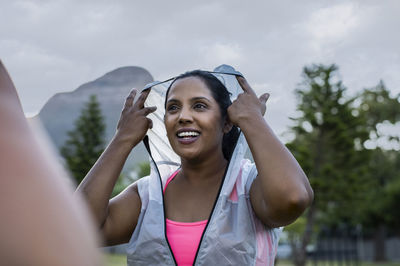 Low angle view of happy woman wearing hooded jacket while standing against sky in park