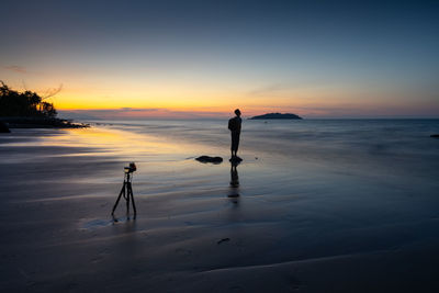 Camera on tripod with silhouette man standing at beach against sky during sunset