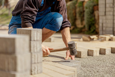 Young man laying gray concrete paving slabs in house courtyard on gravel foundation base.