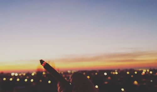Cropped hand holding illuminated cigarette against city during sunset
