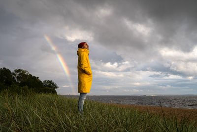 Man in raincoat wear red hat standing on the beach in rainy weather, looks at dramatic cloudy sky
