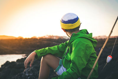 Boy sitting by fishing rod on rock against sky during sunset