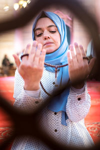 Young woman holding beads while praying in mosque seen through window fence