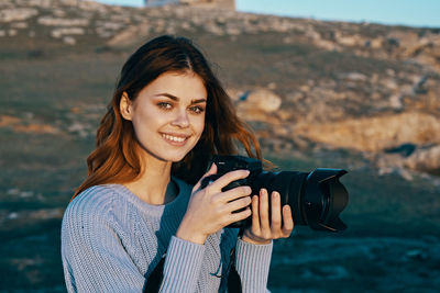 Portrait of smiling young woman holding camera while standing outdoors
