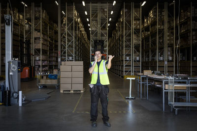 Full length of young male warehouse worker gesturing while using walkie-talkie at storage room