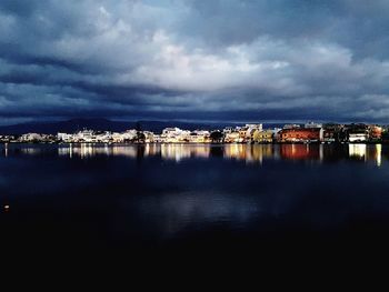 Illuminated cityscape by sea against dramatic sky