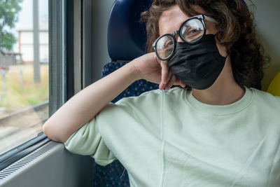 View of young woman traveling by train during pandemic wearing glasses and black mask.