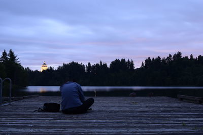Rear view of man sitting on lake against sky at dusk