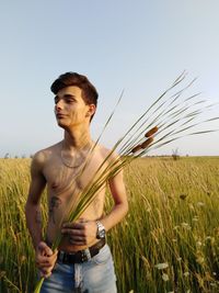 Shirtless teenage boy holding plants while standing on field against clear sky
