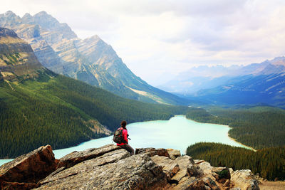 Rear view of man sitting on rock by lake against mountains