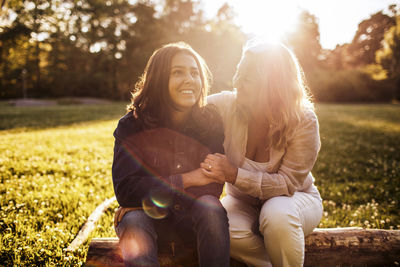 Mother looking at smiling daughter while sitting on log at park during sunny day