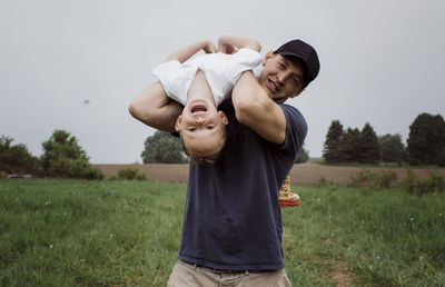 Portrait of happy father picking son upside down while standing on grassy field against sky at park