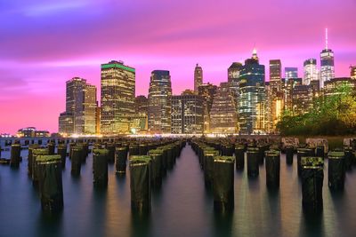 Wooden posts in river against modern buildings at night