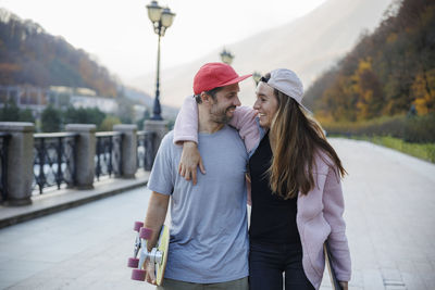 Carefree couple looking at each other walking with skateboards on footpath