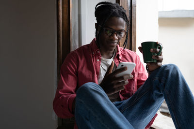 Relaxed charismatic african american man student sits on windowsill with mug of tea and mobile phone