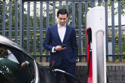 Businessman using smart phone standing at electric vehicle charging station