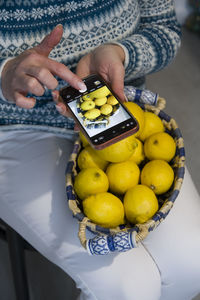 A woman in a blue sweater takes a picture of a basket with ripe yellow lemons,