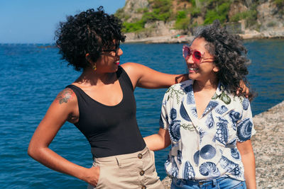 Cheerful tattooed black woman embracing happy mature ethnic friend while looking at camera against sea and mount in sunlight