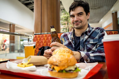 Man eating fast food burger and drinking beer alone in the open area of a restaurant