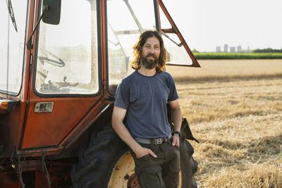 Smiling farmer with hands in pockets leaning on tractor at farm