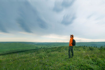 Side view of male hiker with backpack standing on grassy field against cloudy sky