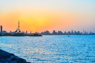 Sea by city buildings against sky during sunset