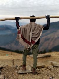 Rear view of man standing with log on mountain peak against sky