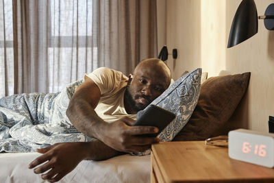 Young man checking smartphone lying on bed at home