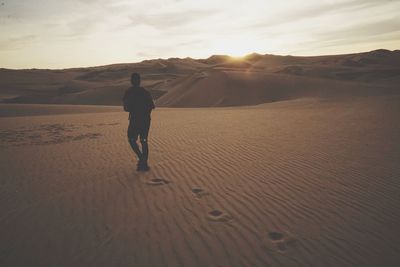 Rear view of man walking on desert against sky during sunset