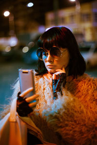 Fashionable woman wearing warm clothing while using smart phone in city at night