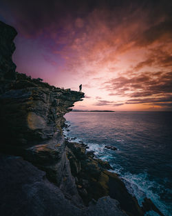 Silhouette person standing on cliff over sea against sky during sunset