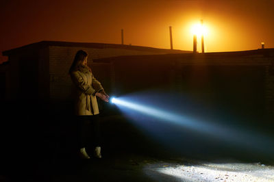 Side view of young woman holding flashlight while standing on road against orange sky during sunset