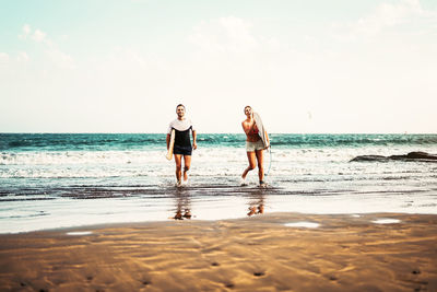 Full length of couple with surfboard walking on beach against sky