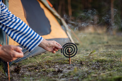 Woman installing burning mosquito repellent coil with smoke on dry tree twig in forest near tent