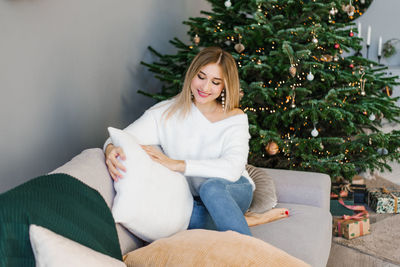 Lovey woman is sitting on a gray sofa near the christmas tree in the living room and holding
