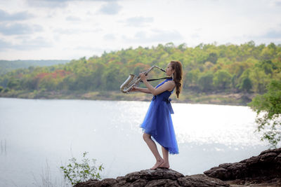 Side view of beautiful young woman in dress playing trumpet while standing on rock by lake