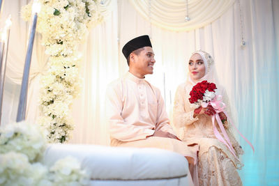 Bride holding flower bouquet by groom sitting at home