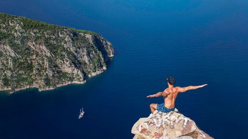 High angle view of shirtless young man with arms outstretched sitting on mountain over sea during sunny day