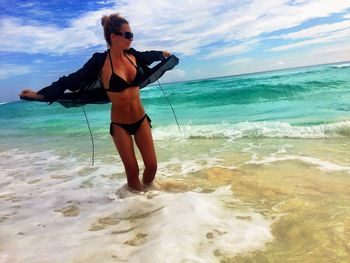 Seductive woman in bikini removing jacket in sea against sky