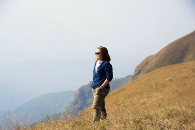 Woman with hands in pocket standing on hill against sky during sunny day