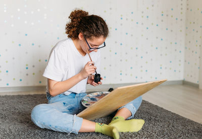 Curly hair girl teenager in glasses in white t-shirt sitting on floor painting on wooden desk 