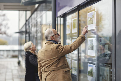 Senior man pointing in glass window by female partner in city during winter